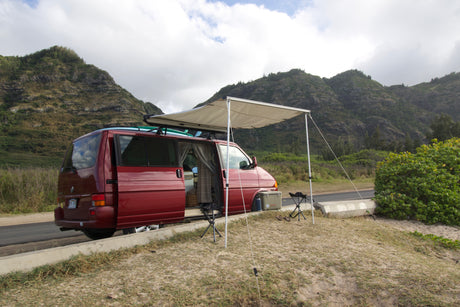 Red camper van with awning & surfboards on a scenic Hawaiian roadside. Perfect for your next RV rental adventure.