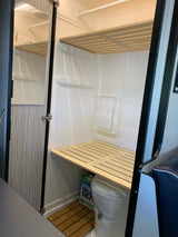 This RV rental features a functional, compact interior with a toilet, tiered slatted shelves, and a wooden floor mat.