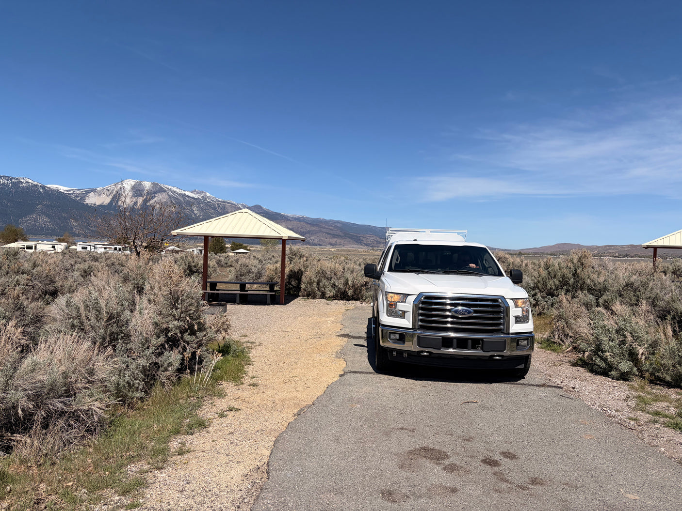 White truck at a scenic RV campground. Distant RVs, snow-capped mountains, and a picnic shelter. Ideal for an RV rental.