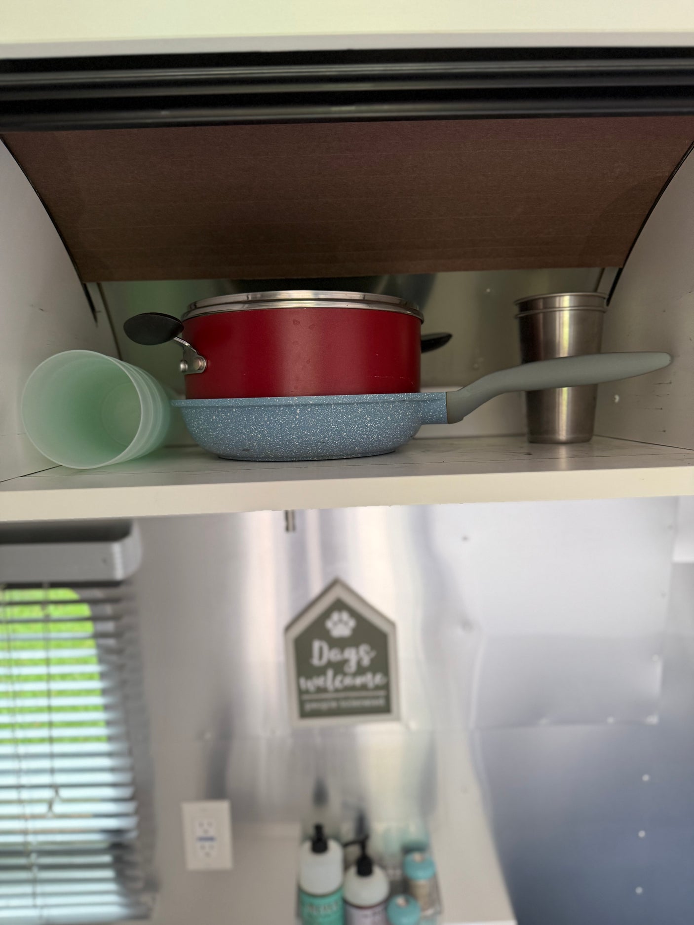 An rv rental kitchen with a red pot, blue pan, and cups neatly stored on a shelf. Ready for compact adventures.
