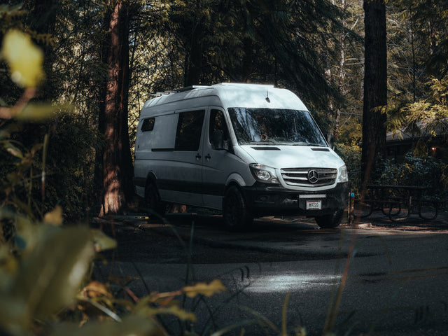 A white Mercedes Sprinter camper van is parked among tall trees