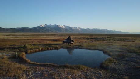 RV rental: A truck with a pop-up tent camps by a tranquil hot spring, reflecting snowy mountains and vast plains.