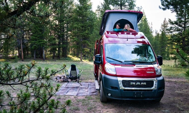 Two people enjoy an RV rental with a pop-top roof at a scenic forest campsite, complete with a fire pit and chair.