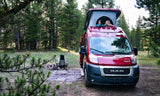Two people enjoy an RV rental with a pop-top roof at a scenic forest campsite, complete with a fire pit and chair.
