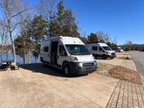 Three white camper vans, possibly RV rentals, parked on a paved waterfront site. One van's side door is open, revealing its interior.