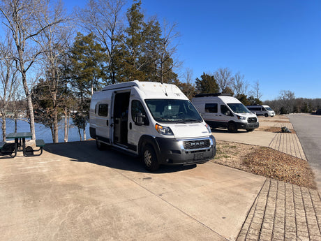 Three white camper vans, possibly RV rentals, parked on a paved waterfront site. One van's side door is open, revealing its interior.