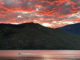 Vibrant red sunset sky over a lake with green mountains. A white boat speeds across the water. Perfect for an RV rental trip!