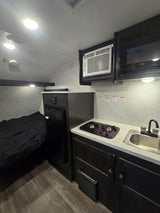 Inside an RV rental: features a bed, compact kitchen with sink/stove, fridge, microwave, and AC unit.