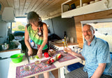 Inside a roomy RV rental, a couple cooks and dines, enjoying wine and picturesque views from their comfortable travel space.