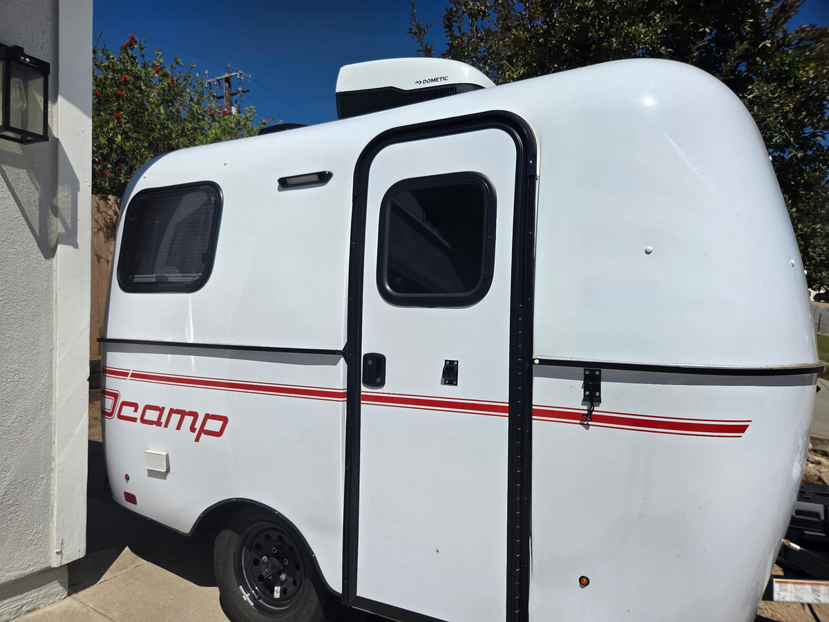  door open & ready for adventure.|This compact white JOCAMP teardrop trailer with red stripes and a Dometic AC unit is ready for an rv rental adventure.|Cozy RV rental interior with a light grey bed
