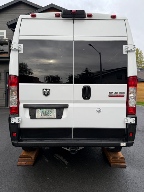 Rear view of a white Ram ProMaster rv rental van, identified by its Alaska "VAN02" plate, parked on wooden leveling blocks.