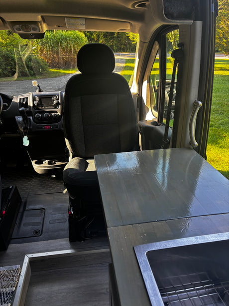View inside an rv rental camper van: driver's seat, dashboard, and kitchen counter with sink. Green outdoors.