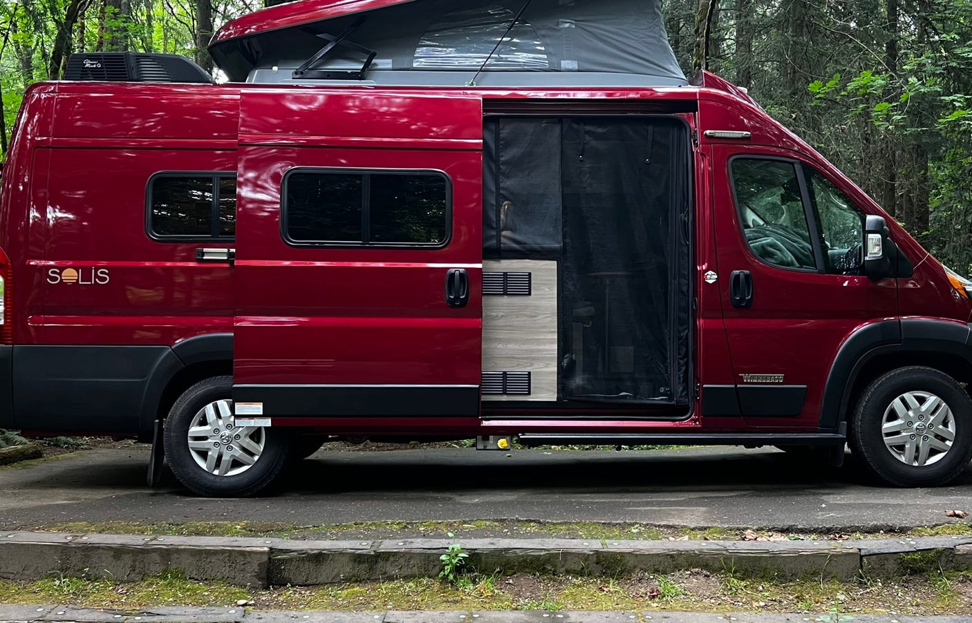 A bright red Winnebago Solis campervan, perfect for an RV rental, showcasing its open side door in a forest.