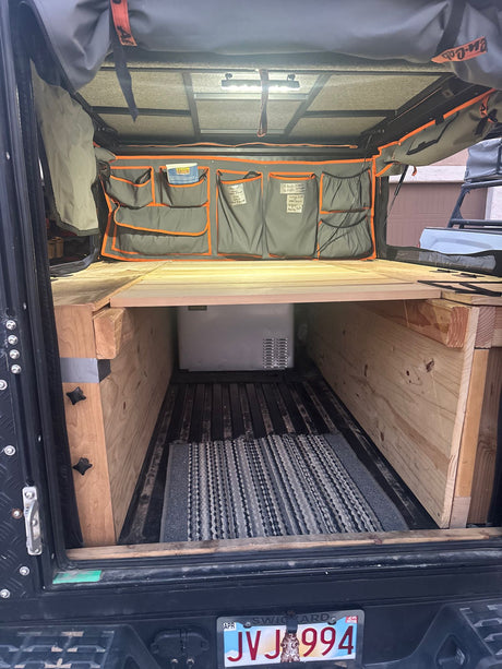 Interior view of a DIY truck bed camper build, ideal for RV rental. Features a custom wooden platform, portable fridge, and storage organizers.
