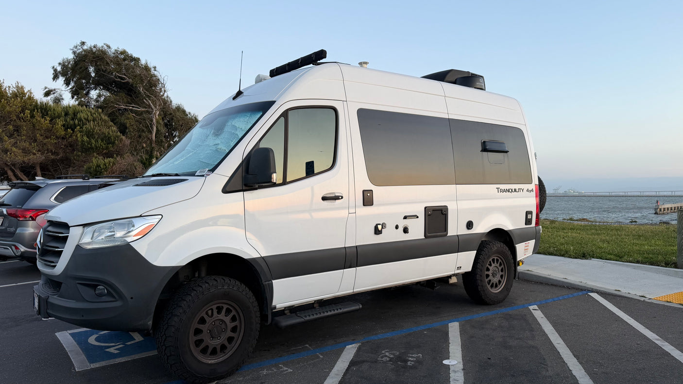 White Mercedes Sprinter rv rental camper van, "Tranquility 4x4," with off-road tires, parked by the water.