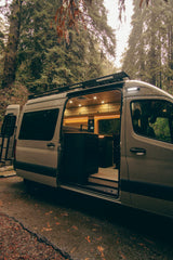 Modern camper van, perfect for an rv rental adventure, parked in a lush forest with its illuminated interior visible.