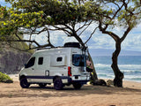 A white Winnebago Revel rv rental camper van sits on a sandy beach under a tree, overlooking ocean waves. Ideal for coastal adventures.