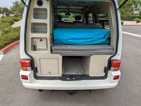 Rear view of a compact camper van's interior, showing folded bed, storage, and sink. Perfect for your next RV rental.
