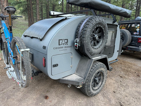 Gray Cedar Ridge teardrop camper RV rental with mountain bike, solar panel, spare tire, and awning ready for forest adventures.