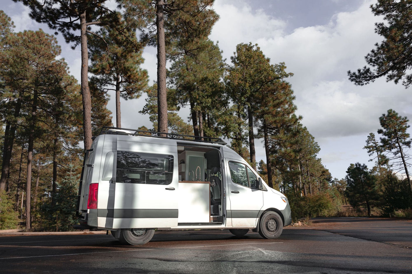 White camper van, side door open revealing its interior kitchenette, parked in a pine forest. Perfect for RV rental.