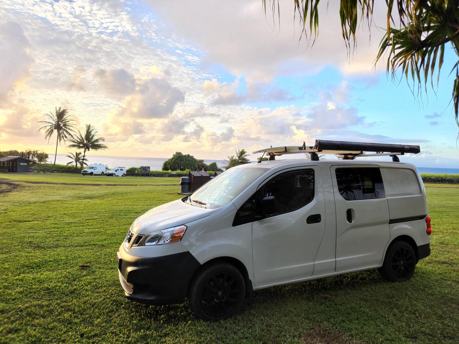 White camper van (likely rv rental) with surfboard and awning parked in a grassy field by the ocean at sunset.