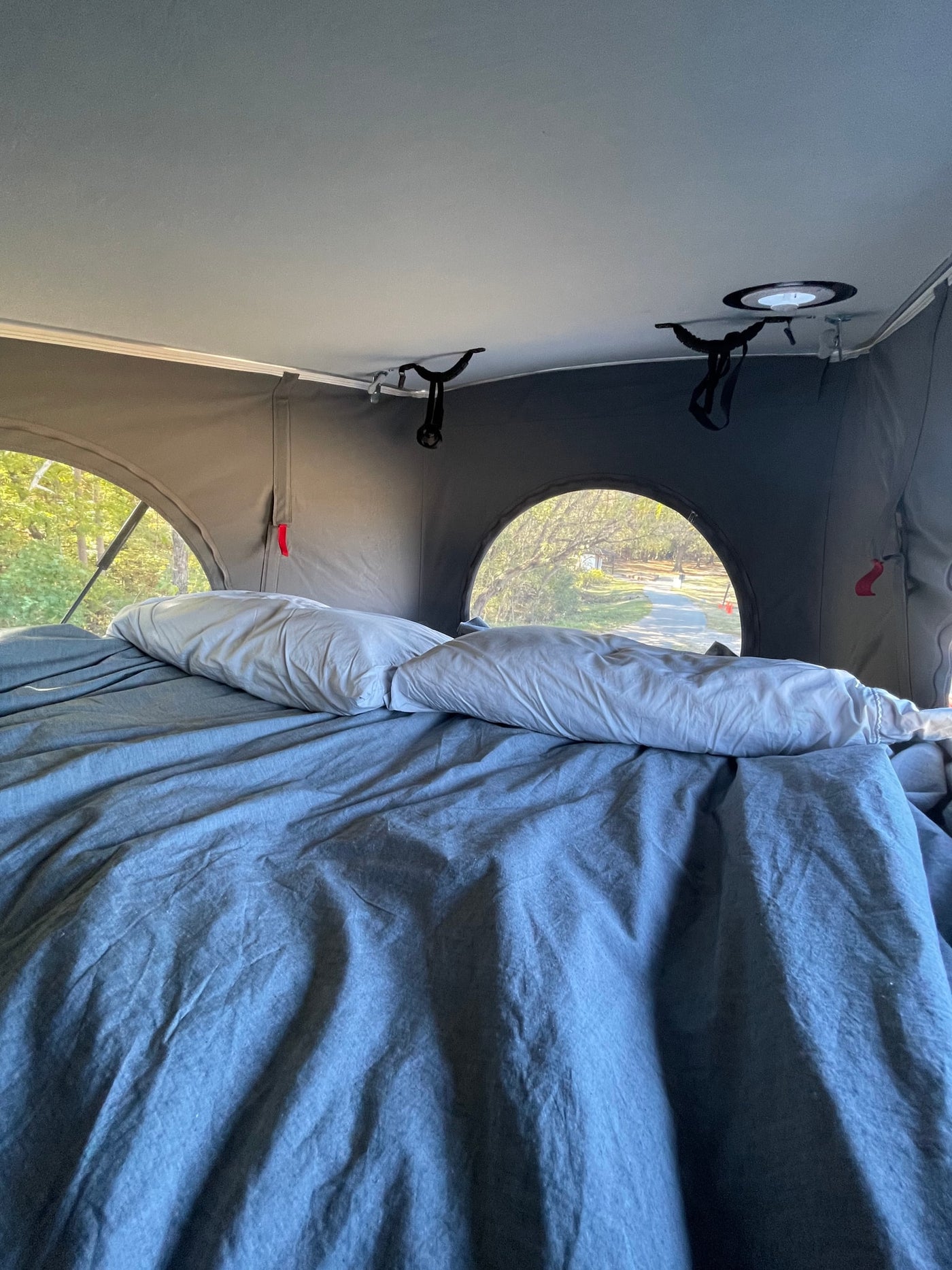 An RV rental interior shows a comfy bed with grey sheets & pillows, looking out arched windows to a wooded path. — photo 14