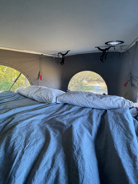 An RV rental interior shows a comfy bed with grey sheets & pillows, looking out arched windows to a wooded path.