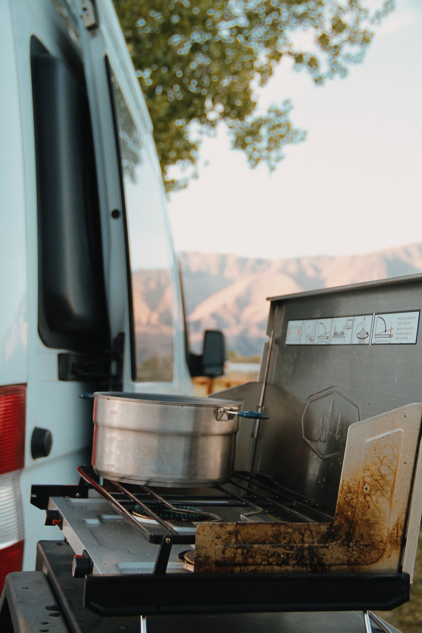A dirty portable camping stove with a pot sits beside an RV rental, ready for outdoor cooking amidst mountains.