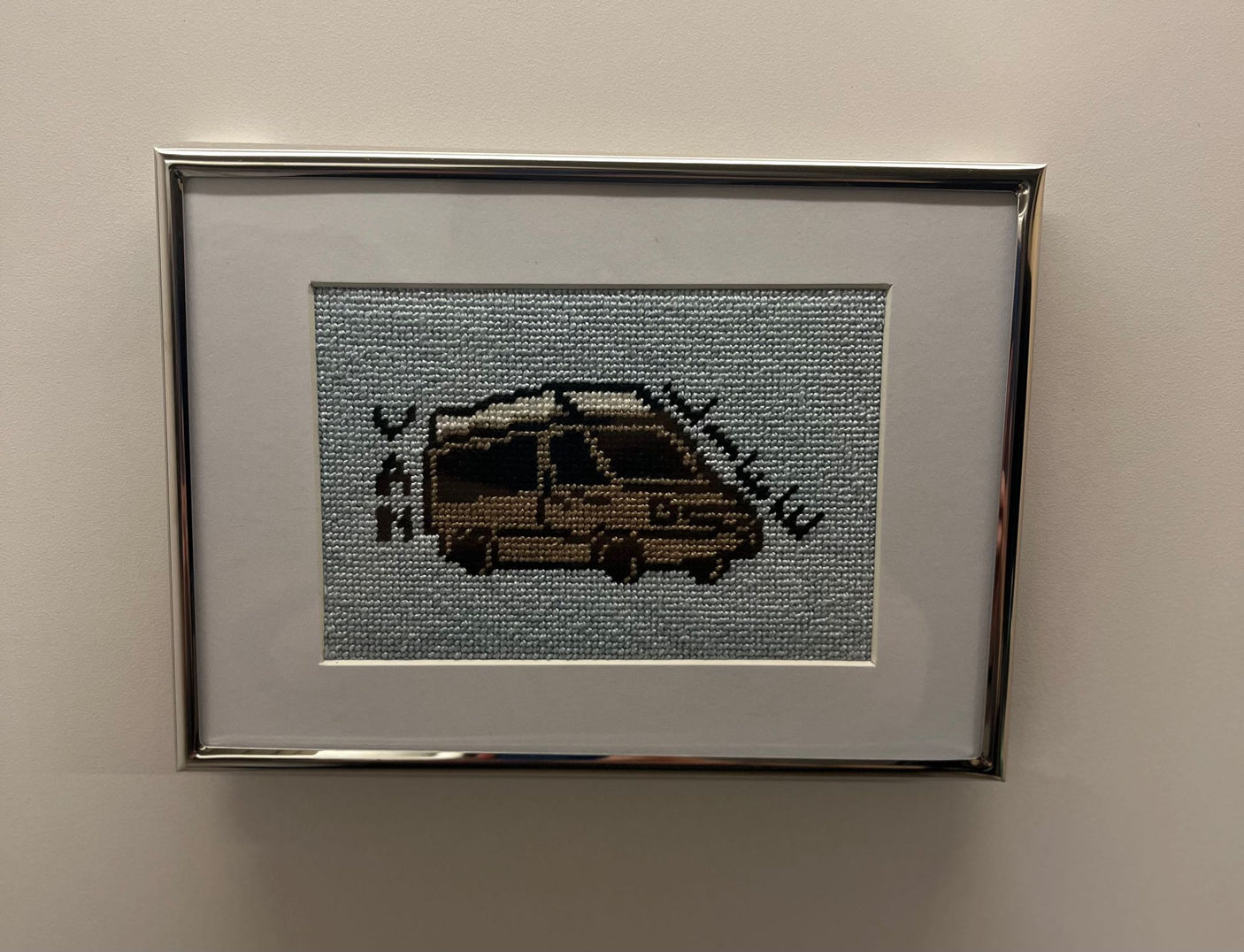 Close-up of a framed cross-stitch artwork, depicting a brown RV with roof details on light blue fabric, ideal for RV rental enthusiasts.