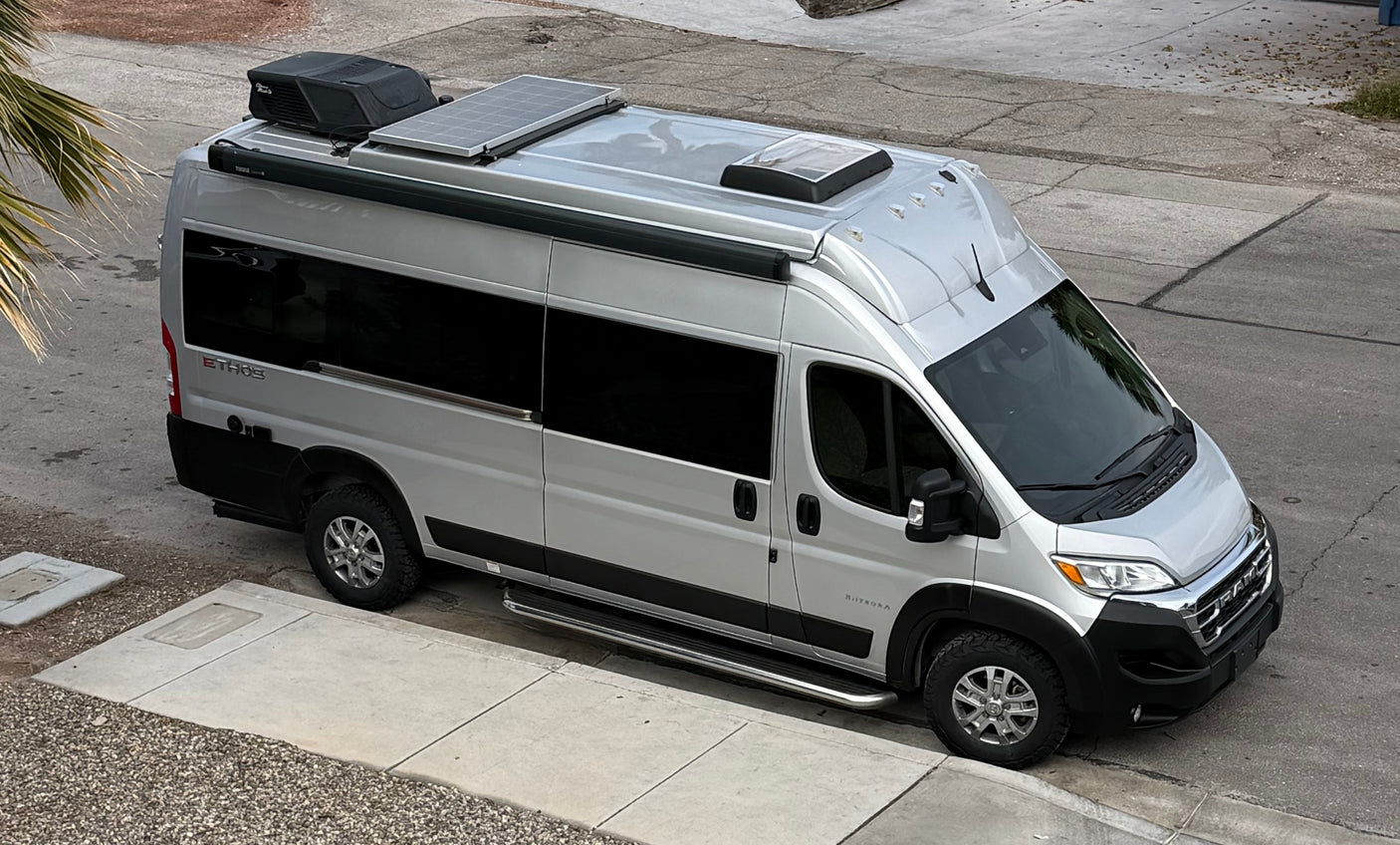 High-angle shot of a silver Ethos RV rental camper van with rooftop solar panels, AC, and all-terrain tires, parked on street.