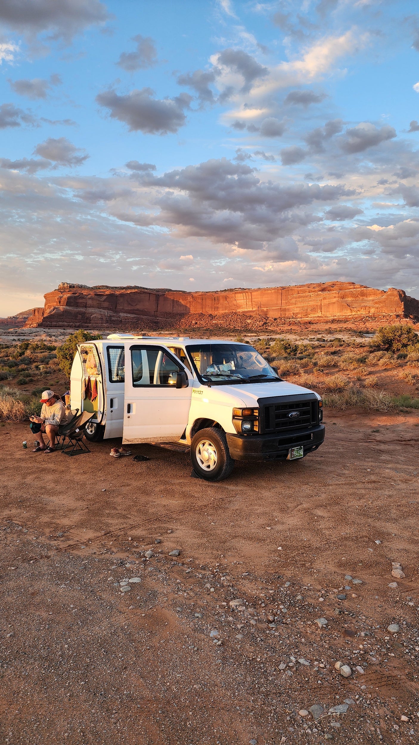 An RV rental Ford van is parked in a scenic red rock desert at sunset, with two people relaxing outside.