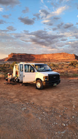 An RV rental Ford van is parked in a scenic red rock desert at sunset, with two people relaxing outside.