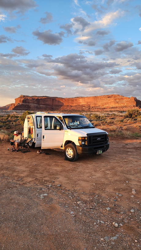 An RV rental Ford van is parked in a scenic red rock desert at sunset, with two people relaxing outside.