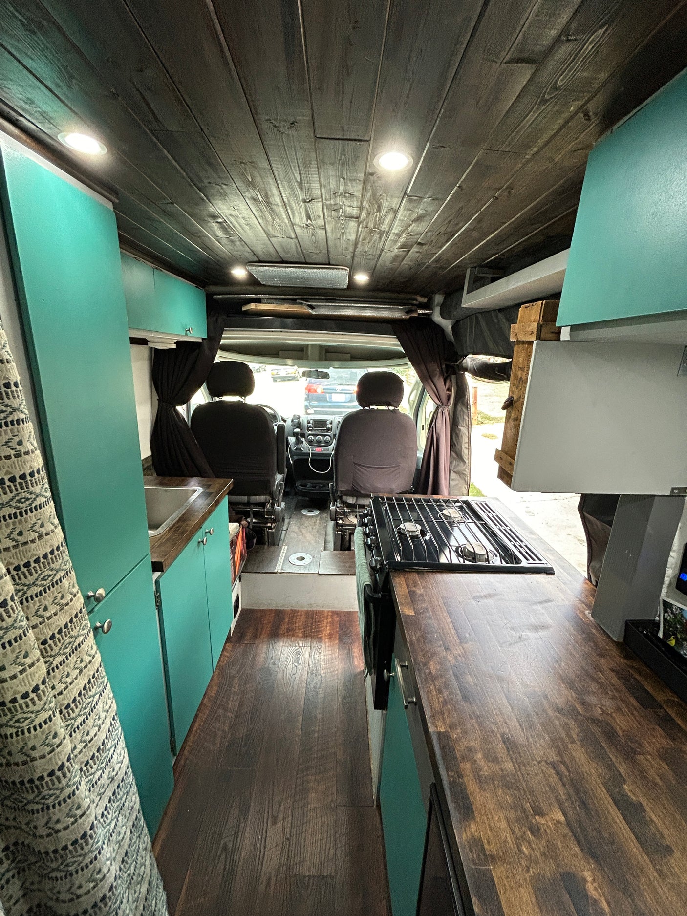 Modern rv rental camper van interior: wood ceiling, teal cabinets, kitchen with sink & stove, leading to front seats.