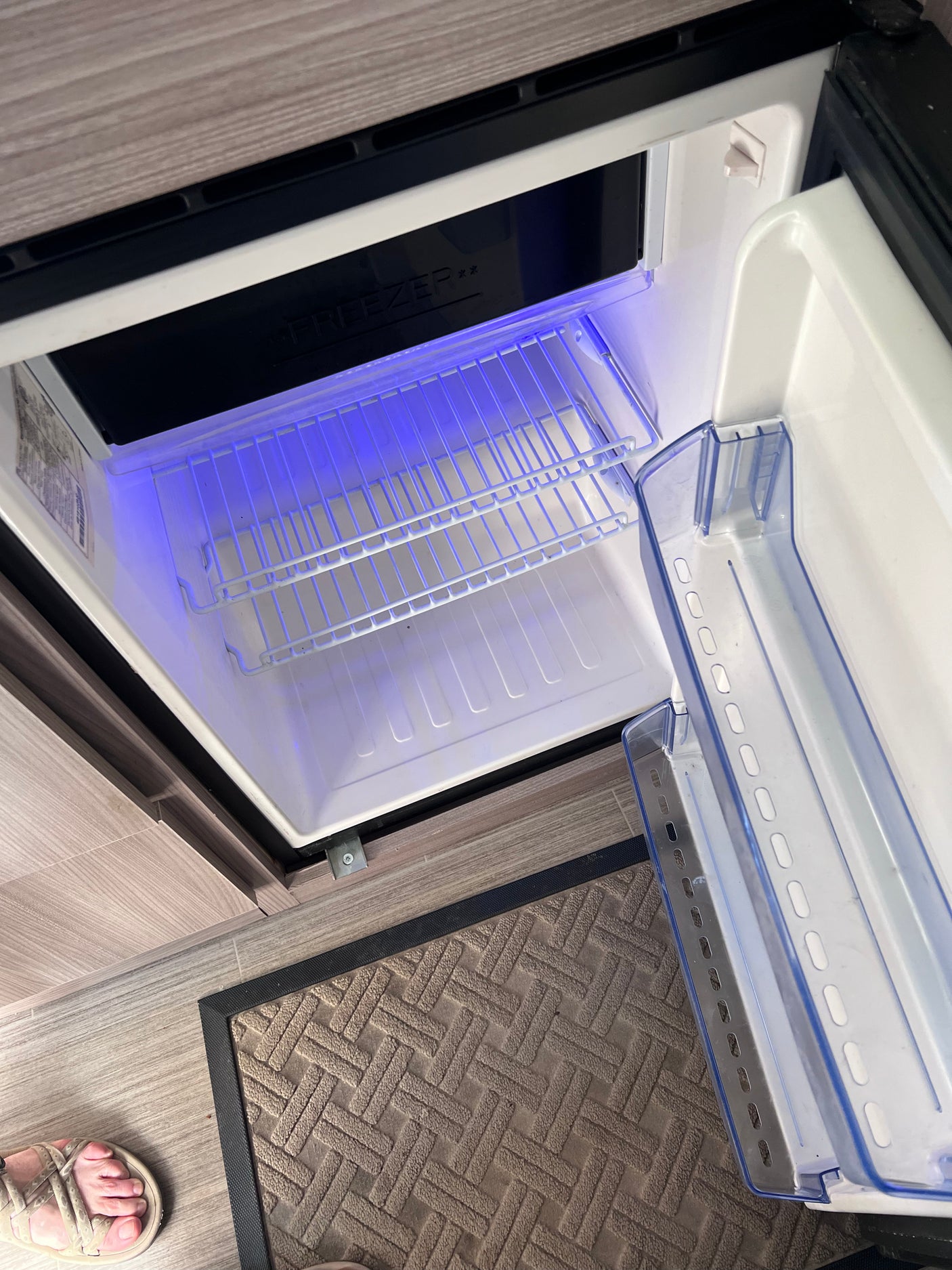 Inside an open, lit rv rental mini-fridge with wire shelves and plastic door bins. A foot in a sandal on a mat.