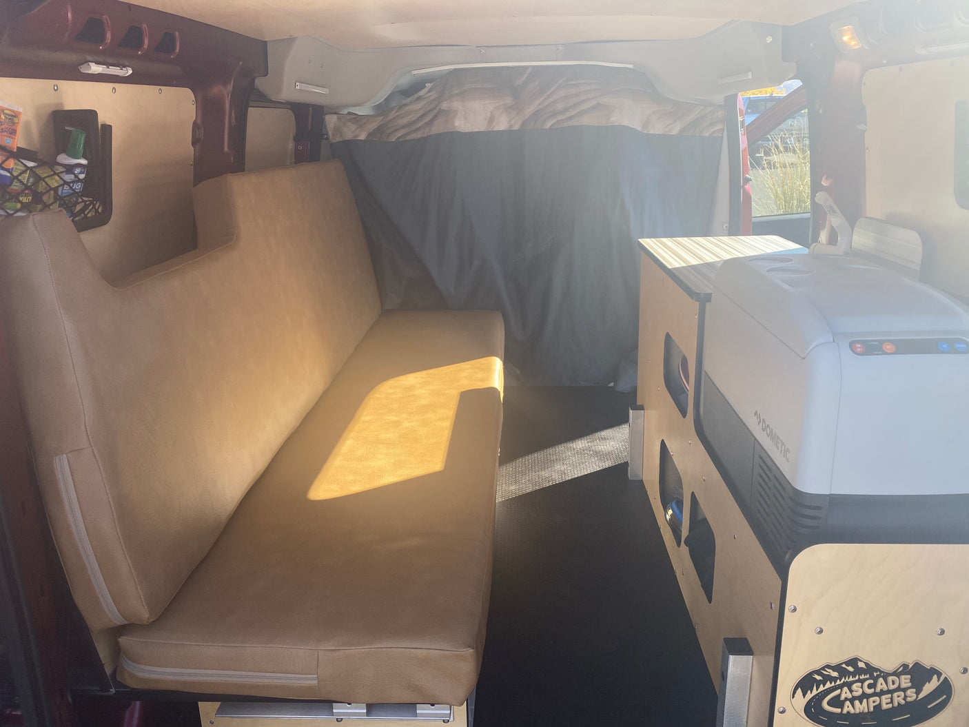 An rv rental camper van interior with a long beige bench, Dometic fridge, and "Cascade Campers" cabinet, ready for adventure.