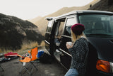 Scenic mountain camping with an RV rental. A woman sips coffee beside her black camper van and outdoor gear.