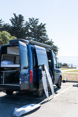 An RV rental van with open doors, surfboards leaning against it, parked at a coastal overlook.