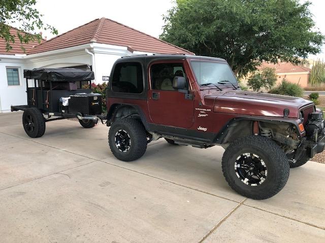 This camping trailer with a red rooftop tent & tarp is an excellent RV rental option for forest adventures.|This black off-road camping trailer with a tent frame is parked in a grassy forest. Great for your next RV rental adventure.|Red Yakima rooftop tent on a trailer — photo 6