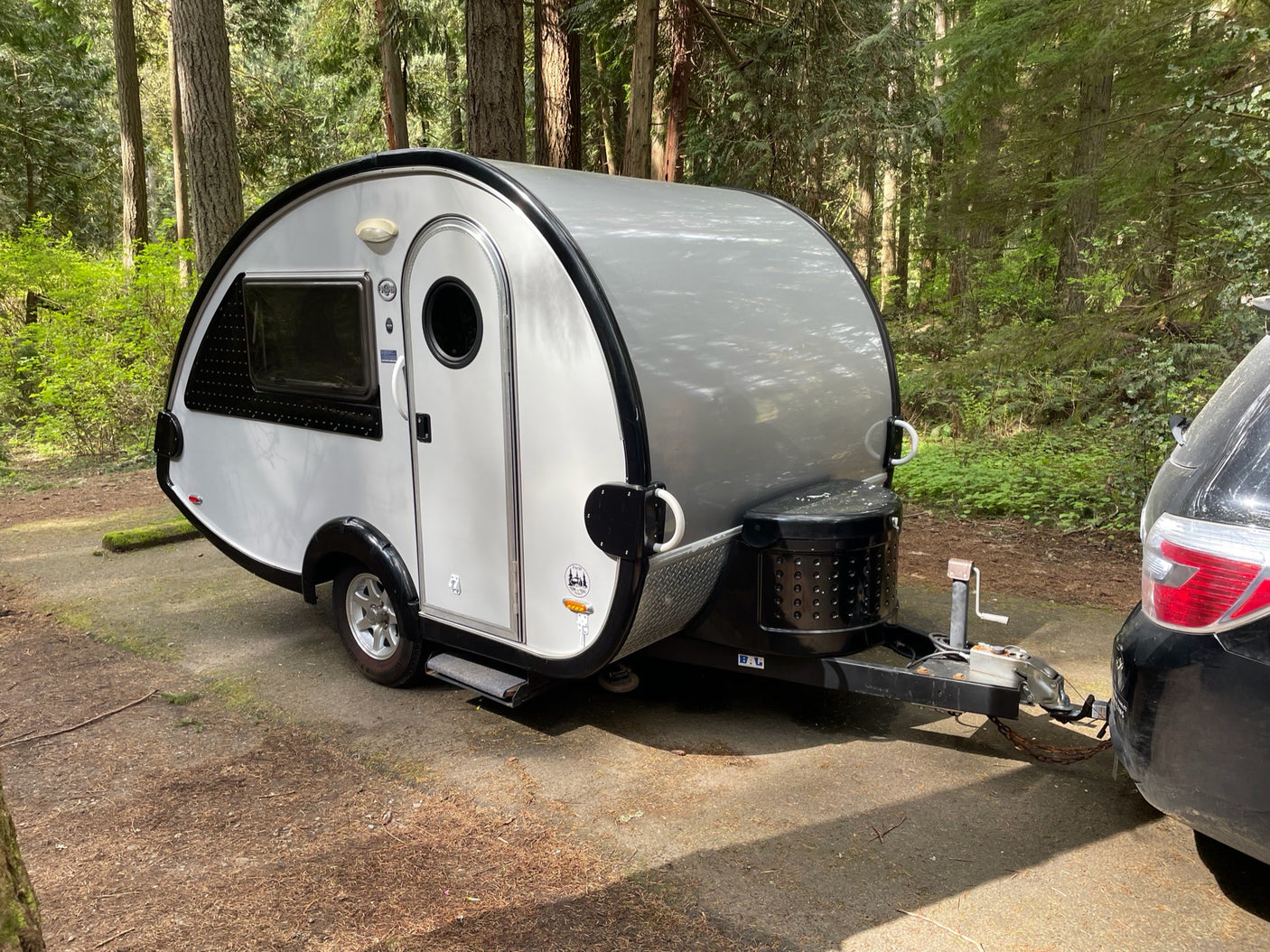 Small teardrop RV rental parked in a peaceful forest campsite, hitched to a car. Ideal for outdoor adventures.