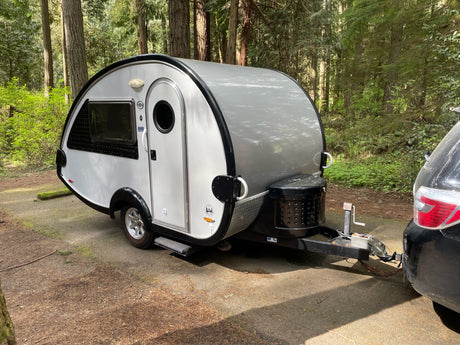 Small teardrop RV rental parked in a peaceful forest campsite, hitched to a car. Ideal for outdoor adventures.