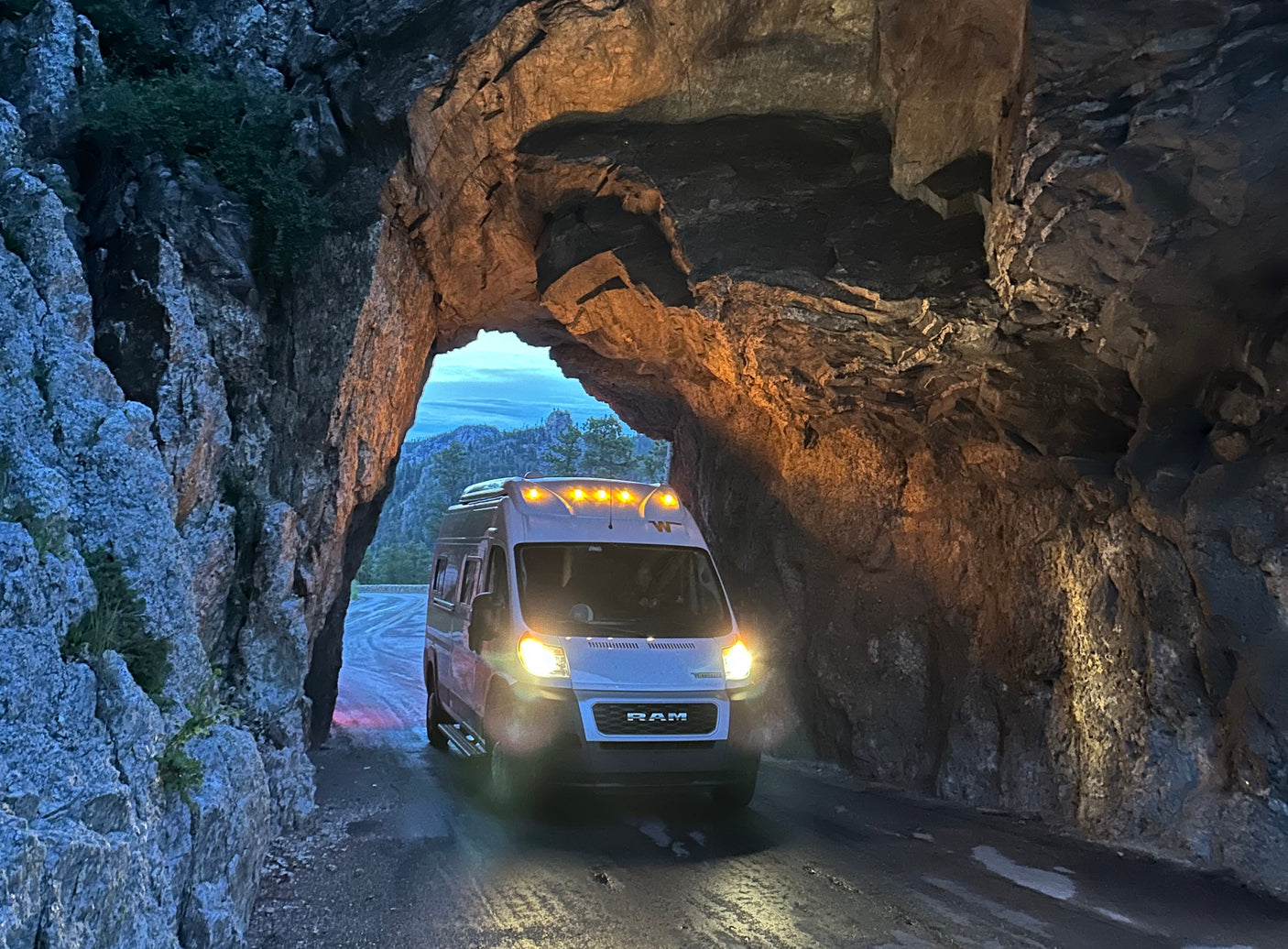 An RV rental emerges from a natural rock tunnel. Its bright headlights illuminate the rugged passage and a mountain view.