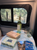 Inside an RV rental: wine, glasses, cribbage, card, & dice games, plus a tic-tac-toe set, all set against a beautiful forest & river view.