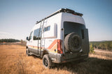 Rugged white Storyteller Overland adventure van with ladder, roof rack, and rear spare tire, ideal for an rv rental outdoor escape.