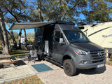 A modern gray Sprinter camper van rv rental with awning extended, off-road tires, and outdoor setup at an RV park.
