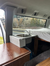 Inside an RV rental: a compact camper van kitchenette with a ZUHNÉ sink and faucet, next to a cozy bed.