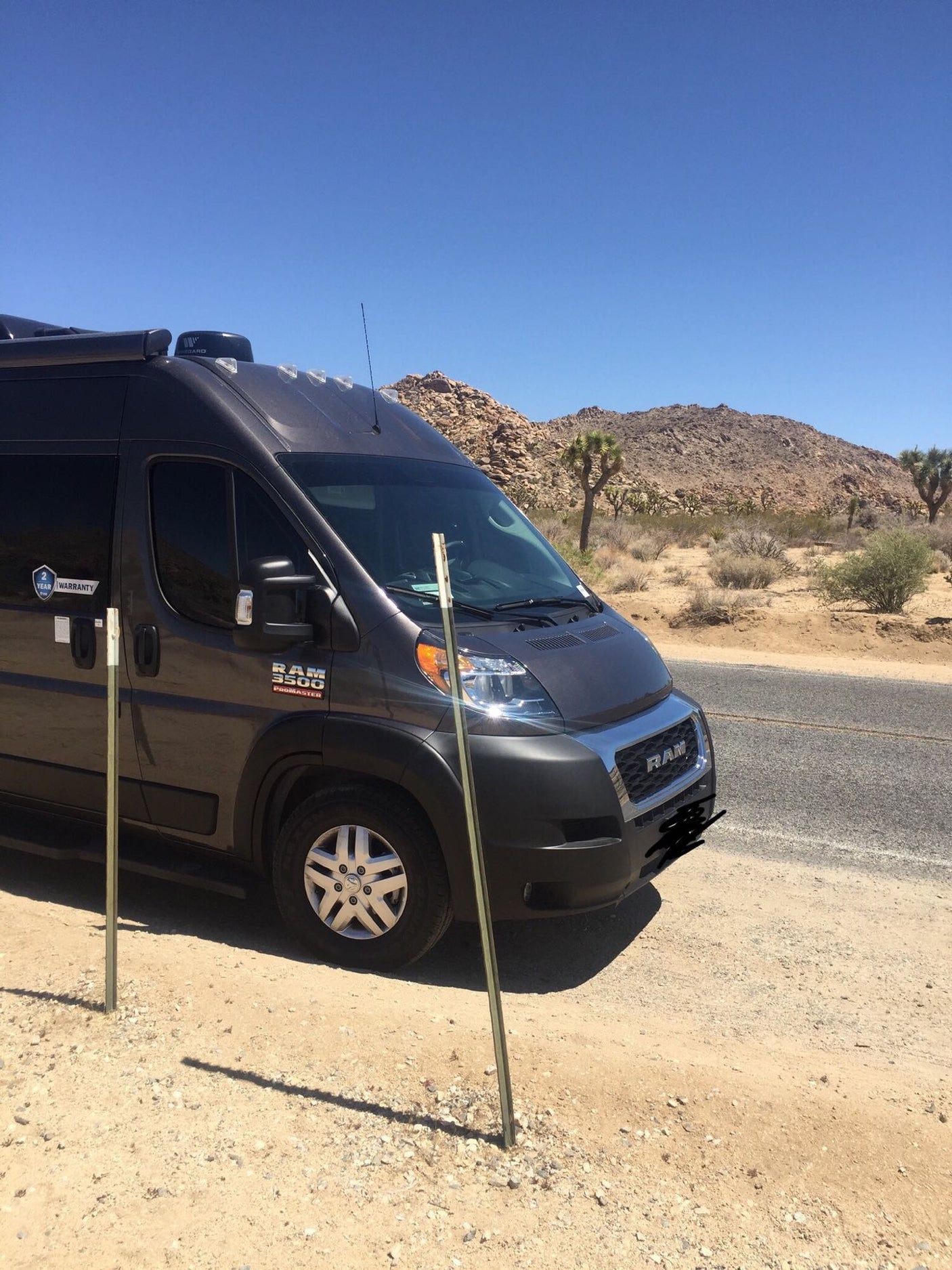A dark grey Ram ProMaster rv rental campervan parked on a dirt shoulder in a sunny desert with Joshua trees.