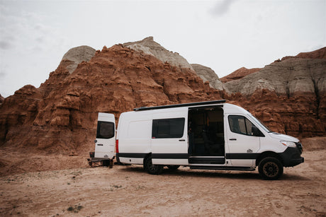 A white RV rental van with open doors is set up for camping amidst dramatic red and grey desert formations.