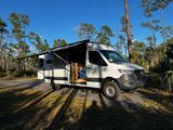 RV rental adventure: a white Mercedes Sprinter camper van with an awning extended at a sunny campsite among tall trees.