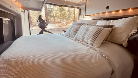 Cozy RV rental interior: bed with pillows facing a snowy forest view & bike outside. Ideal for travel.